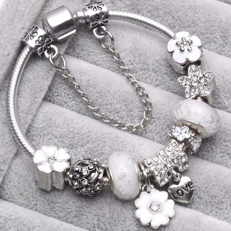 A silver-colored jewelry bracelet with multiple charms including floral designs and a cross, displayed on a grey background.